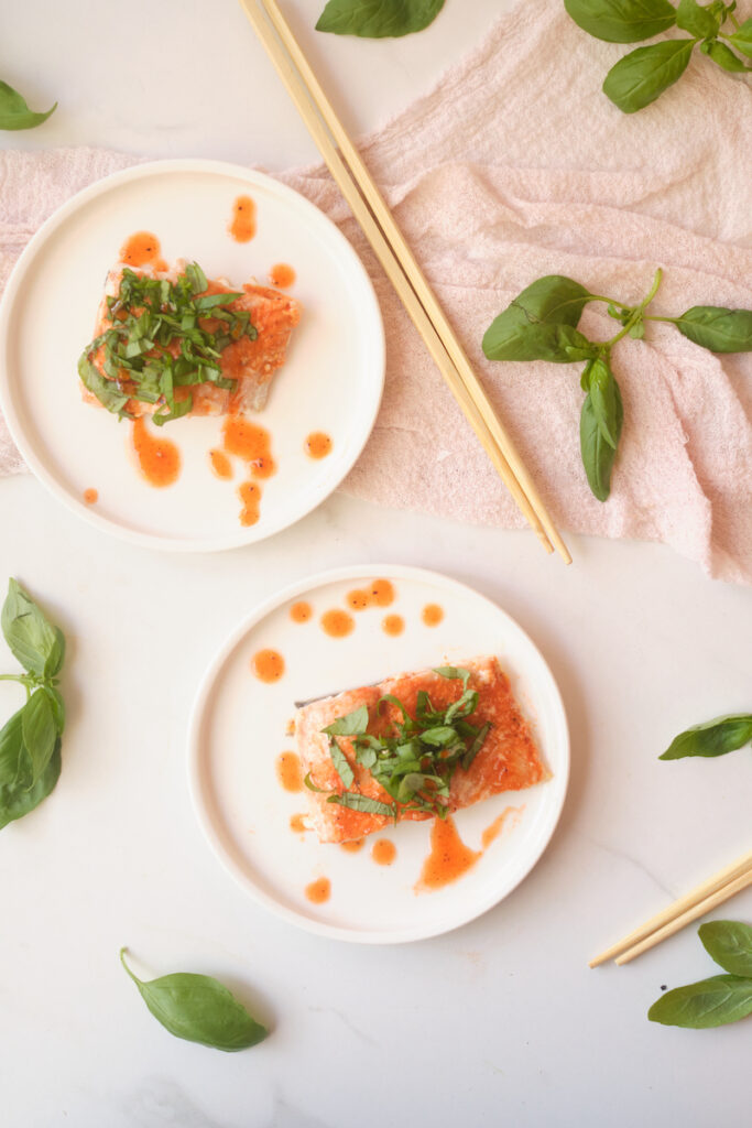 Birds eye view of honey sriracha salmon fillets with basil, plated with basil leaves strewn about and chopsticks in view