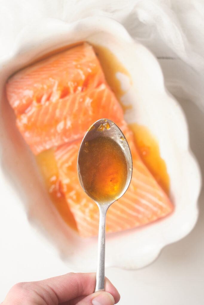 A hand holds a spoon full of apricot glaze over a dish containing fresh salmon fillet