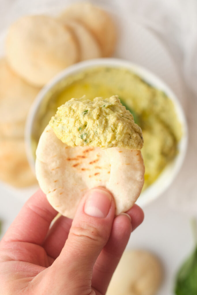 A woman's hand holds pita bread dipped in basil avocado hummus dip