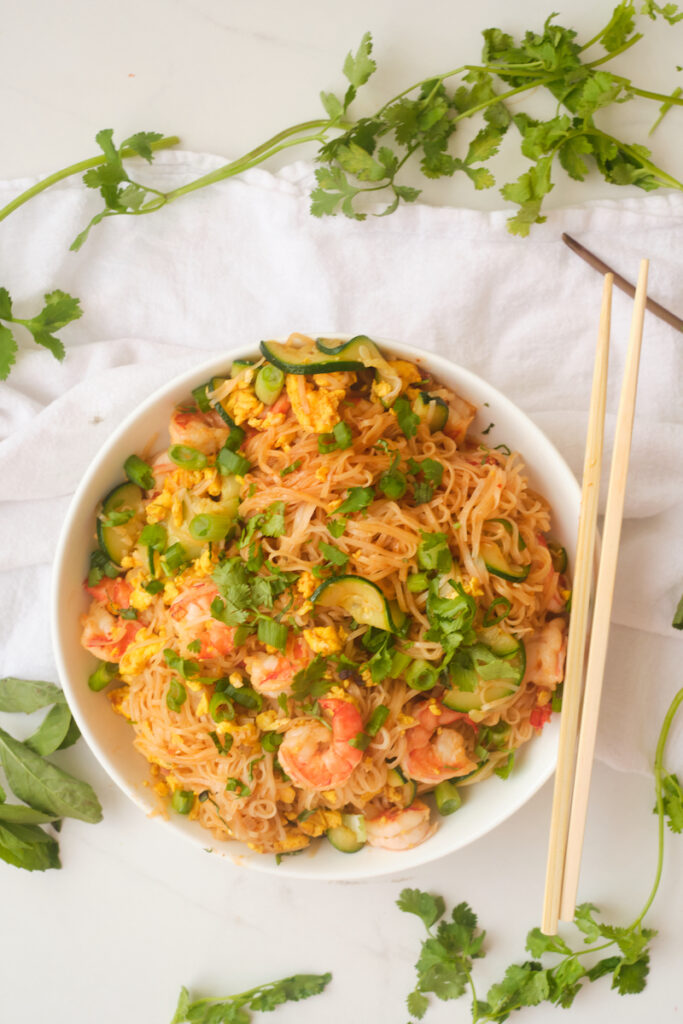 Spicy shrimp noodles in a bowl with chopsticks and thai basil and herbs surrounding it