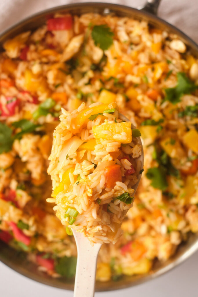 A wooden spoon with a mouthful of thai pineapple fried rice with basil above the frying pan