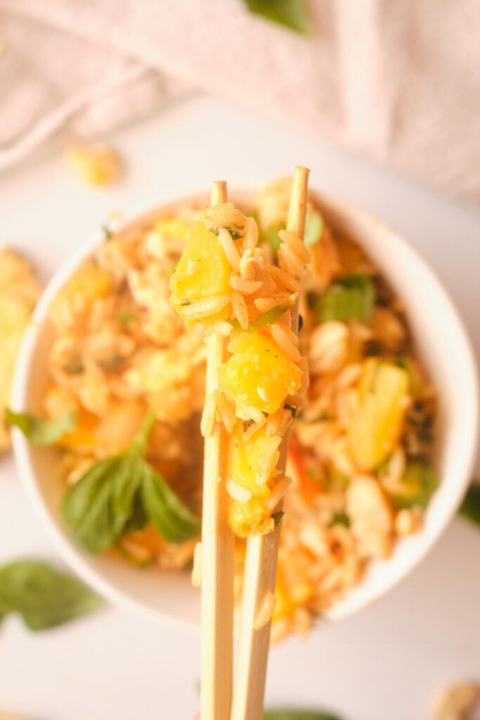 Chopsticks holding a bite of thai pineapple fried rice above a bowl
