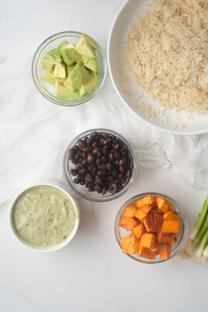 Roasted sweet potato, black beans, basil yoghurt dressing, avocado, green onions and rice separated into dishes to show ingredients for a sweet potato buddha bowl