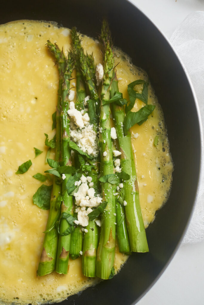 Process shot showing an asparagus omelette being made, with asparagus spears, goat cheese and basil placed on a partially cooked omelet in the pan