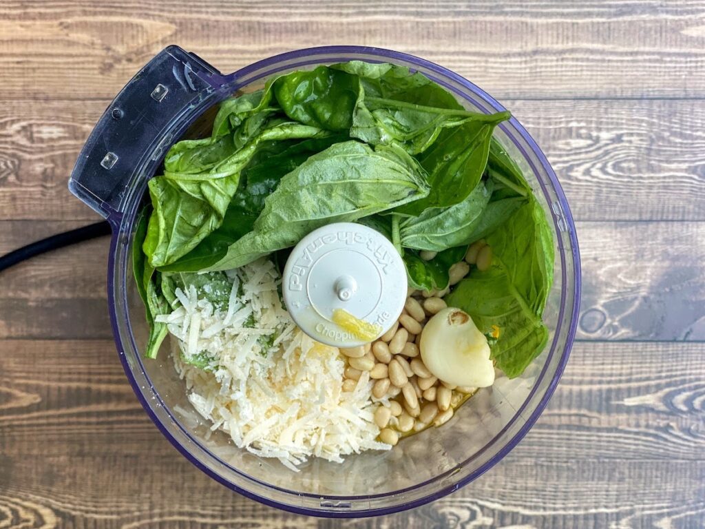Ingredients for Tuscany basil pesto in a food processor including tuscany basil leaves, garlic, pecorino and parmesan cheese, pine nuts and olive oil
