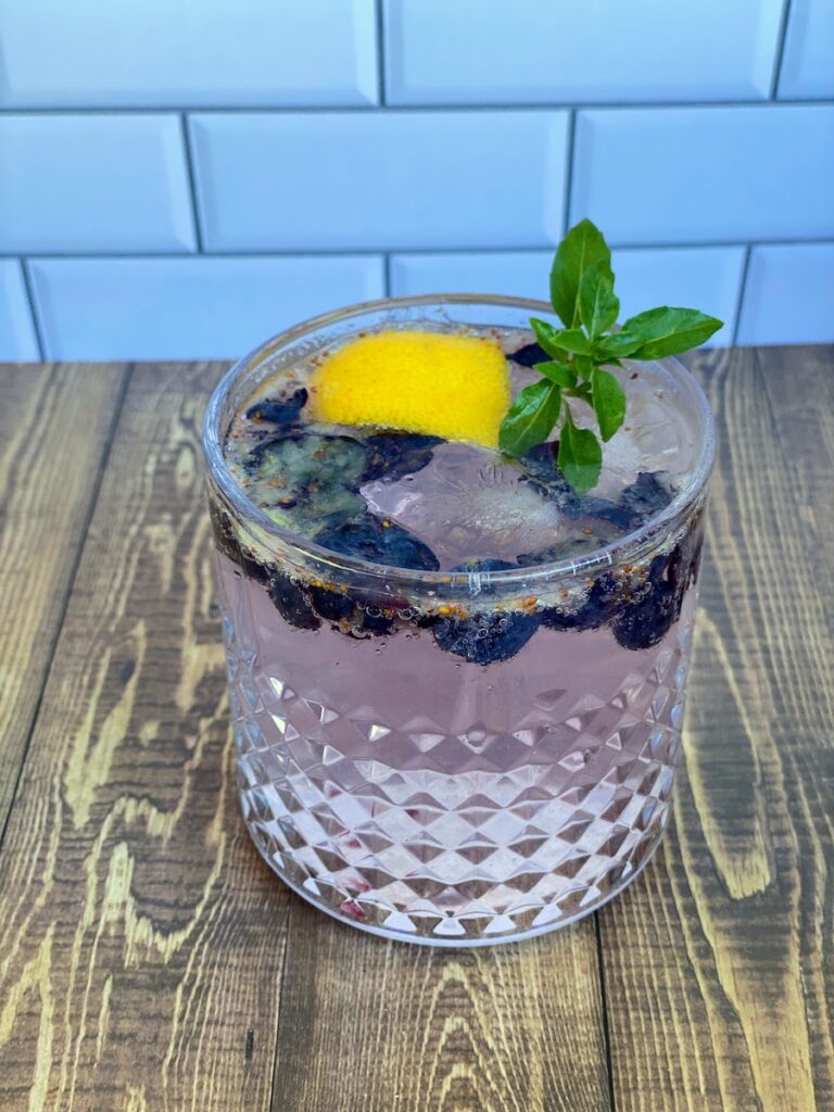A lemon basil mocktail made with lemon basil simple syrup, fresh lemon and blueberries, a sprig of spicy bush basil, and sparkling water