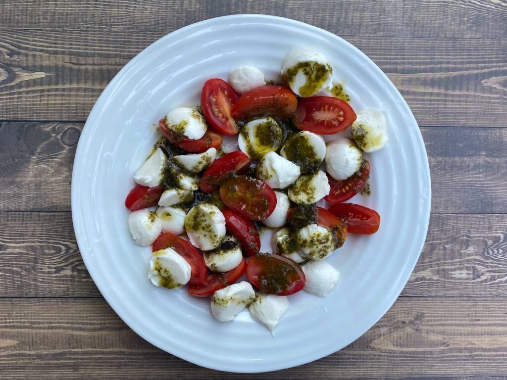 A twist on a caprese salad made with tomatoes, bocconcini and basil stem salad dressing. Plated on a white plate.
